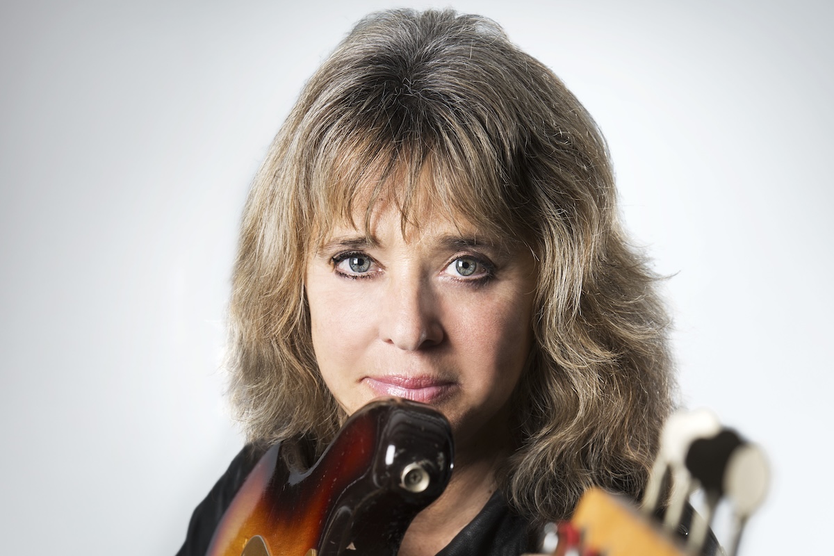 Rock icon Suzi Quatro posing for a close-up studio portrait while holding her electric bass guitar against a light background.